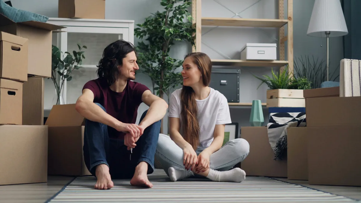 Couple sitting on the floor of an empty room, surrounded by moving boxes, smiling